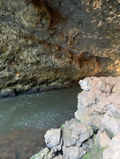 The image depicts a narrow body of water, likely a stream or river, flowing between rocky walls. The rocks are jagged and irregular, with varying shades of grey and brown, and some patches of green moss. The water is murky, with some surface ripples and bubbles visible. The location is Mururi, Kenya.