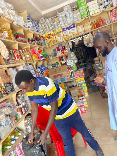 Inside a retail shop in Samaru, Nigeria, two adult males are present amidst shelves heavily stocked with various consumer goods. In the foreground, a male with short dark hair, wearing a horizontally striped polo shirt (blue, yellow, white) and dark blue trousers, is bent over a red plastic chair, actively reaching into a black plastic bag. To the right and slightly behind him, a bald, bearded male, dressed in a light-colored traditional garment, stands upright, looking down at and operating a smartphone held in his left hand. The shop walls are lined with multiple wooden shelves displaying an assortment of products including cooking oils, cereals (Kellogg's Corn Flakes, Golden Morn), packaged snacks (Munch Cheesy Stix, Indomie noodles), personal care items, cleaning supplies, and bulk goods like toilet paper and flour/sugar bags. Bunches of garlic hang from the ceiling in mesh bags. The floor is visible as a light-colored surface, and the overall environment is well-lit.