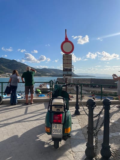 This image depicts a vibrant coastal scene in Cefalù, Italy, specifically from a promenade overlooking the Lungomare beach and the historic port area.
In the immediate foreground, a dark green Piaggio Vespa scooter is parked, facing away from the viewer. Its license plate, "PA 11 0868," is clearly visible, and it features a beige seat and a rear rack. To the right of the scooter, a series of dark, ornate metal bollards connected by chains serve as a barrier. Behind the scooter, a tall metal pole supports a large circular red and white "no access" or "do not enter" sign at its apex, with a red fabric strip hanging from the top. Below this sign, several rectangular informational signs with black text and symbols on a white background are stacked vertically.
In the midground, to the left, two individuals stand leaning against a railing, observing the sea. The male, with gray hair, wears a dark green t-shirt and khaki shorts, and has his right hand raised to his forehead as he looks towards the horizon. The female, beside him, wears a light-colored short-sleeved top and dark blue jeans, carrying a dark bag over her left shoulder. To the far right, a partially visible person is holding a camera or smartphone, seemingly capturing the view.
The midground transitions to a sandy beach and the clear, turquoise water of the Mediterranean Sea. Numerous people are scattered across the beach, sunbathing, and swimming in the water. A blue beach umbrella is visible on the sand. A stone pier or breakwater extends into the sea from the right, with several people visible on it. Paddle boats, colored white and blue, are docked near the shore on the left.
The background features lush, green, tree-covered mountains and hills that descend towards the coastline, framing the bay. Distant buildings indicate a town along the far side of the bay. The sky is bright blue with scattered white cumulus clouds. Strong sunlight casts distinct shadows of the bollards and the scooter onto the light-colored, block-paved promenade.