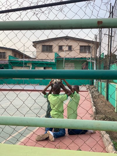 Three young boys are playing with a soccer ball in a basketball court in Akure, Nigeria. They are kneeling and holding the ball above their heads. The court is surrounded by a chain-link fence and green walls. The fence is visible in the foreground, and the court surface is red and lined with white. Two of the boys are wearing green shirts, while the third is wearing a light blue shirt. The image is taken from a slightly elevated position behind the fence. The boys are interacting and laughing, appearing to be having fun while playing. The scene is located in a residential area, with a building in the background. The building is a two-story structure with a flat roof and several windows. There are also some trees and bushes around the court. The sky is clear and blue. The image is likely taken in the daytime, and the lighting is good. The image is in focus and sharp.