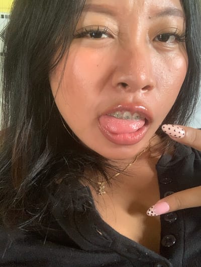 The image is a close-up selfie of a young woman with dark hair and braces. She is wearing a black top with a low neckline and a gold necklace. Her tongue is sticking out, and her nails are painted pink with black polka dots. The image appears to have been taken indoors, as suggested by the lighting and the background, which is out of focus and shows a wall with a patterned border. There is no indication of the specific location within Jajag, Indonesia.