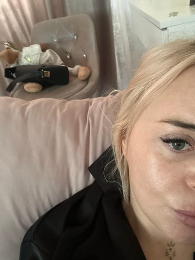 A close-up, rotated image features a woman with blonde hair, fair skin, and light-colored eyes. She has a small tattoo near her mouth. The image is centered on her face. She is lying on a light pink pillow. She is wearing a black garment or top. Beside the pillow is a small, ornate chair with a light-colored cushion, accented with rhinestones. A doll is seated on the chair, also with fair skin. The doll is wearing a white outfit. A small black bag with gold hardware is positioned to the side of the doll. Part of a white piece of furniture is visible in the upper left corner. There is nothing in the image to indicate the city of Высокий, Russia.