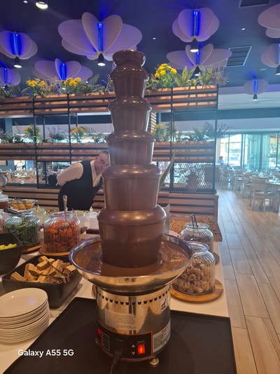 An indoor scene captured in a modern dining or event space, likely a hotel or buffet in Gouves Municipal Unit, Greece, features a prominent chocolate fountain dessert station. The image is rotated 90 degrees clockwise.

At the center, a large, multi-tiered chocolate fountain, made of stainless steel and brown plastic, circulates melted chocolate. To its right, an array of clear glass jars holds various toppings, including what appear to be dried figs, dried apricots, and other dried fruits, along with possibly nuts or wafers. A black tray with serving tongs holds more wafers, and a stack of white plates is ready for use. The base of the fountain has an orange "Switch" button and "Power" text, along with other labels such as "MADE IN CHINA," "DO NOT IMMERSE," and "READ INSTRU."

Behind the chocolate fountain, a man dressed in a white shirt and black vest, presumably a staff member, stands attentively.

The background features decorative elements and other parts of the establishment. The wall on the left is adorned with multiple white, flower-shaped light fixtures, each emanating a blue glow from its center. Below these, there are small circular spotlights. Further back, wooden slatted partitions are visible, interspersed with green and yellow foliage, creating a natural, inviting atmosphere. Beyond these partitions, a dining area with numerous white tables and chairs is dimly visible, along with large windows letting in ambient light.

The bottom right corner of the image displays the watermark "Galaxy A55 5G," indicating the device used to capture the photo.