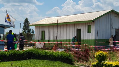 This daytime image, taken in Tchere, Democratic Republic of the Congo, depicts a scene likely associated with a humanitarian or public health operation. The focal point is a simple, white wooden building with a green-painted base and trim, and a corrugated metal roof. Its walls feature vertical planks, and it has several green-framed windows and doors.

In the foreground, a manicured lawn with green and yellow-green bushes creates a clear division. An area in front of the building is cordoned off by red and white striped barrier tape and orange mesh fencing, indicating a controlled or organized space.

Several people are visible within and around this fenced area. To the left, near a partial tent structure, two individuals are prominent: one in a blue shirt and dark pants, and another wearing a high-visibility yellow vest over a dark top. A third person in a patterned shirt stands nearby, close to a green bin. Above them, a white flag proudly displays the text "THE ALLIANCE INTERNATIONAL" and "ALIMA," with other partial words like "GNER" and "VER" also visible.

Towards the right side of the building, a person dressed in green scrubs and blue gloves is actively engaged with a yellow container or bag near one of the doors, suggesting a task related to health, sanitation, or supplies. Further right, a few other individuals, including one in a light-colored long-sleeved shirt and a hat, are standing near another doorway.

The scene is under a bright, sunny sky with scattered white clouds. The overall atmosphere appears functional and organized. The bottom right corner of the image features the watermark "Joseph Kisuki."