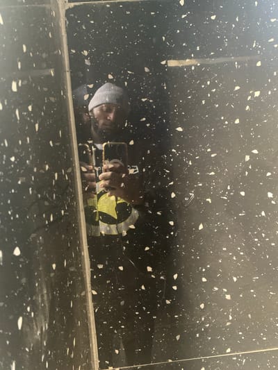 The media file shows a reflection on a dark, speckled surface, likely a countertop or polished floor. The reflection depicts a person from the chest up, wearing a blue and white striped hooded garment. The person appears to be holding a smartphone with a gold frame, and their hands are visible, reaching towards the phone. A yellow circular logo or design is also reflected on the surface, partially obscured by the person's hands. The scene suggests the person is using their phone, possibly taking a picture or video, given the way they are holding it. The lighting is somewhat dim, and the reflection creates a sense of depth on the speckled surface. There is no visible text within the frame of the reflection itself. The location context provided is Rukuba Road, Nigeria, though this information is not directly inferable from the visual content alone.