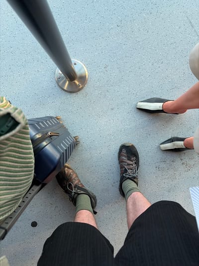 This media file shows a high-angle view of the lower legs and feet of several individuals standing on a speckled gray floor. There are two people on the left wearing black and white mules, and two people on the right wearing athletic shoes and socks. One of the individuals on the right is wearing black patterned shorts, and the other is wearing dark shorts and a green and white striped garment that might be a bag or clothing. A blue suitcase with silver accents is positioned in the foreground, near the feet of the person wearing the striped garment. A metal railing with a circular mounting plate is visible on the left side of the frame. The setting appears to be indoors, possibly a waiting area, given the presence of the railing and suitcase. The lighting suggests it is daytime. No text is visible in the image.