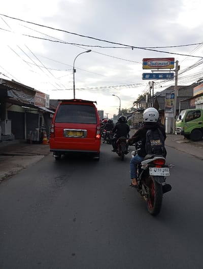 The image shows a street scene in the city of Kutawaringin, Indonesia, located at latitude -6.972 and longitude 107.532. A person wearing a white helmet and a black jacket with a backpack is riding a white motorcycle on a paved street. The motorcycle has a black license plate with the letters "D 4193 JBE" and the numbers "08-20". The person is riding the motorcycle toward the camera. There are other motorcycles in the background. There is a red van parked on the right side of the street. The van has a black license plate with the letters "D 1883 XE". There are buildings on both sides of the street. There is a sign for "Indomaret" on the right side of the street, indicating a local convenience store. The sky is overcast.