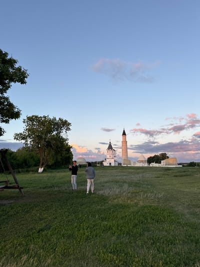This image captures an outdoor scene in Bolgar, Russia, likely during the late afternoon or early evening given the soft, warm lighting and the colors in the sky. The sky is a pale blue with wispy clouds tinged with pink and orange, indicating sunset or sunrise.

The foreground is dominated by a vast expanse of green grass, which appears somewhat overgrown and uneven. In the mid-ground, two individuals are present. One person, wearing a gray hoodie and light-colored pants, is standing, possibly looking out at the scenery. Another person, in a dark top and gray pants, is bent over, holding a smartphone and appearing to be taking a picture or video. They are positioned on the grassy area.

Further in the background, several architectural structures are visible, characteristic of Bolgar's historical sites. There's a tall, slender tower with a dark pointed roof, resembling a minaret. Adjacent to it is a white building with a dome and a cross on top, suggesting a religious structure like a church. The buildings are constructed with what appears to be stone or brick and have a historic appearance. A large, mature tree with a white-painted trunk stands between the grassy area and the buildings, adding a natural element to the scene.

The overall atmosphere is serene and contemplative, with the individuals seemingly enjoying the views and the ambiance of the historic location as the day winds down. No text is visible in the image.