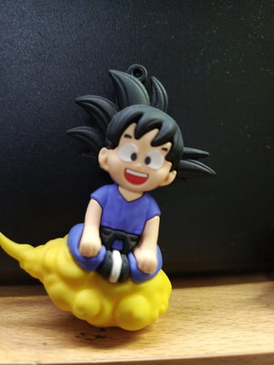 The image displays a chibi-style figurine of the character Goku from the Dragon Ball series. Goku is depicted with his signature black spiky hair, light skin, and a joyful, wide-open-mouthed smile. He wears a purple short-sleeved top, a black belt with a white central stripe, and blue pants. The figurine is seated on a vibrant, bright yellow, fluffy cloud-like object, which represents the Flying Nimbus. A small black loop is visible at the top of Goku's head, suggesting it might be a keychain or hanging ornament.

The figurine is positioned against a dark, textured background, possibly a wall or panel, which appears black or very dark grey. Below the cloud, a warm-toned wooden surface, likely a shelf or table, is visible, showing some wood grain. The lighting appears even, highlighting the figurine's colors and details. There is no visible text in the image. The scene conveys a sense of cheerfulness through Goku's expression. This item is located in Guayaquil, Ecuador.