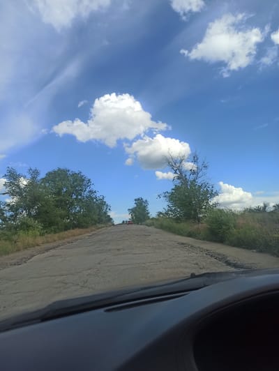 The image depicts a rural roadside scene with a clear blue sky filled with fluffy white clouds. A dirt road runs alongside a grassy verge with trees and vegetation. A portion of a vehicle's interior, specifically the side mirror and part of the dashboard, is visible on the right side of the frame, suggesting the photo was taken from inside a car. The sunlight indicates it is daytime, and the clear sky suggests fair weather. The landscape appears to be open and natural, characteristic of a rural Ukrainian setting. No people or specific events are identifiable, but the composition implies a journey or a pause during travel. The overall mood is peaceful and scenic.