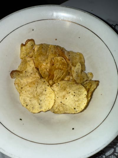 A close-up, top-down shot shows a white ceramic bowl filled with potato chips. The bowl has a thin, gold-colored rim. The potato chips are mostly round or irregularly shaped, with a golden-yellow color and visible seasoning specks. Some chips appear folded or curled. The background is dark and out of focus, suggesting an indoor setting. The image does not provide any specific information about the location in the City of Pematang Siantar, Indonesia.