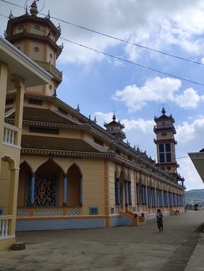 The image captures the exterior of a large, ornate yellow building with a distinct architectural style, likely a temple or pagoda, in Lộc Quý, Vietnam. The structure features multiple tiered roofs adorned with intricate decorative elements and multiple towers, one of which is tall and rectangular with blue window-like panels, while another is octagonal. The building is lined with a colonnade of blue-topped pillars supporting arched openings. Decorative screens and patterned tiles are visible on the facade. The ground in front of the building is paved. A single person, wearing a hat and patterned clothing, is walking away from the camera along the paved area. The sky is blue with scattered white clouds, and power lines are visible overhead. In the background, a hillside with scattered buildings and trees can be observed.