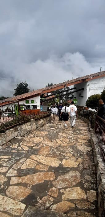 The image depicts a stone-paved pathway at Monserrate sanctuary in Bogotá, Capital District, Colombia, under an overcast sky with significant cloud cover. The pathway, constructed from irregularly shaped, light brown and grey flagstones, slopes gently uphill from the foreground. To the left of the path, a low stone wall supports a planter with sparse vegetation, bordering a white building with a reddish-brown tile roof. This building features green and white signage, suggesting an informational or commercial point. An overhead structure with dark beams extends from this building, providing a covered walkway. To the right, a dark metal railing runs parallel to the pathway, separating it from a lower, indistinct area.

Approximately eight individuals are present on the pathway. Three individuals are prominently visible in the mid-ground, walking away from the camera, uphill. The left-most of these three wears a white jacket and light pants. The central individual wears a dark skirt and a white t-shirt. The right-most individual wears a light-colored t-shirt with a graphic on the back and light-colored pants. Other individuals are further ahead on the path or near the buildings. A partial view of a person in a dark jacket is seen on the far right, behind the metal railing. The environment is characterized by dense fog and clouds obscuring the distant background, with some trees visible behind the structures. The pathway appears damp, possibly from recent precipitation or ambient moisture.