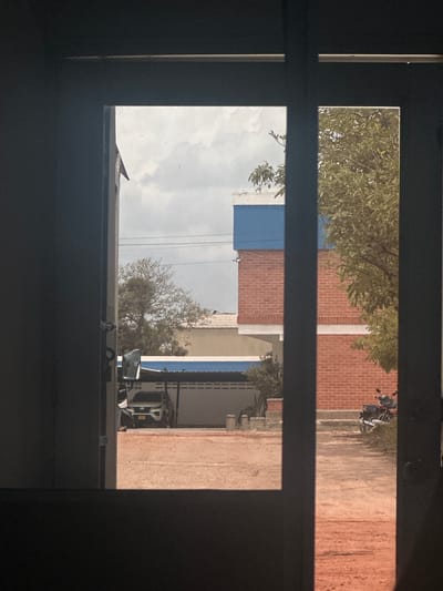 This media file shows a view through a window or door frame, with horizontal bars dividing the scene. The image appears to be taken outdoors, possibly in a rural or semi-rural area given the presence of dirt pathways and buildings with exposed brick.
In the upper portion of the image, a tree with green foliage is visible, along with a brick building and a motorcycle parked on a dirt path. The sky is partly cloudy. The lower portion of the image shows more of the brick building, a blue-colored section, and what looks like a large truck or utility vehicle with visible equipment. A bare tree with thin branches is also present. The overall scene suggests daytime. There is no discernible text in the image. The perspective suggests the viewer is looking out from a shaded interior. The scene is consistent with a location in Juan Mina, Colombia, characterized by its climate and typical structures.
