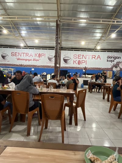 The image depicts the interior of a coffee shop named "Sentra Kopi" in Banda Aceh, Indonesia. The establishment appears to be casual, with a high ceiling supported by metal beams and a corrugated roof. Fluorescent lights illuminate the space. The walls are adorned with a large banner featuring the coffee shop's name, "SENTRA KOPI," in prominent pink lettering, accompanied by illustrations of coffee cups. Below the banner, a patterned mural is visible.
Several tables, predominantly made of wood, are arranged throughout the space, each set with plastic chairs in an orange-brown hue. Patrons are seated at various tables, engaged in conversations or enjoying their beverages. In the foreground, a woman wearing a grey hijab is seated at a table. To her left, another individual is partially visible. In the middle ground, men are seen sitting at tables, some appearing to be in conversation. In the background, more customers and staff can be observed, as well as what appears to be a counter area with shelves stocked with goods, possibly coffee-related products or snacks. The floor is tiled with light-colored squares. A green bowl containing packaged snacks is visible in the lower right corner of the image, placed on a wooden surface. The overall atmosphere suggests a busy, community-oriented coffee shop.