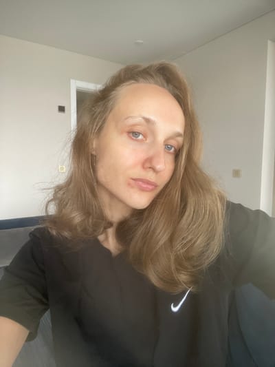 A woman with long, light brown hair and blue eyes is pictured in a medium shot. She is wearing a black t-shirt with a white Nike logo on the left chest. The background is an interior space with light-colored walls and a doorway visible. The woman appears to be indoors, likely in a residential setting. The specific location in the city of Подшивалово, Russia, cannot be determined from the image alone.