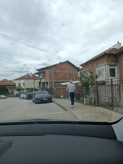 A woman with blonde hair pulled back in a ponytail walks down a sidewalk in Svilengrad, Bulgaria, on a cloudy day. She is wearing a light grey sweatshirt and dark blue sweatpants with a white stripe down the side. She is walking away from the camera. Several parked cars are visible on the street to her left, and houses with red tile roofs line the street on both sides. The foreground shows the dashboard of a car, indicating the image was taken from inside a vehicle.