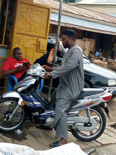 In Arawa, Nigeria, a man wearing a grey thobe and black skullcap sits on a silver and blue motorcycle. The motorcycle is positioned on a wooden surface that appears to be a street or walkway. To the left of the motorcycle, another man in a red t-shirt is seated on a blue plastic chair, looking at his mobile phone. Behind the seated man, a yellow metal structure, possibly a shop or doorway, is visible. Further in the background, a black car is parked, and other structures with corrugated metal roofs suggest an urban environment. The overall scene depicts a street-level view with people and vehicles typical of a Nigerian city.