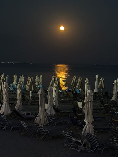 The image depicts a serene night scene at a beach in Nea Kallikratia, Greece, illuminated by a bright, full moon. The moon hangs in the dark sky, casting a warm, golden glow onto the calm sea, creating a shimmering reflection. In the foreground, numerous beach chairs and umbrellas are neatly arranged on the sand, most of them closed and covered, indicating the beach is not actively in use at this hour. The chairs are of various designs, with some featuring striped patterns. The overall atmosphere is peaceful and tranquil, highlighting the natural beauty of the coastal landscape under the moonlight.