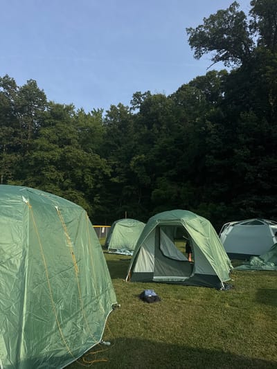 An outdoor scene depicts a campsite with several green tents set up on a grassy field. In the foreground, a large green tent is partially visible on the left. To the right, a smaller, open green tent shows a person's legs inside. Several other tents, one light gray and two more green, are scattered across the field. In the background, a dense line of trees forms a backdrop against a clear blue sky. A yellow baseball backstop is visible behind some of the tents. The ground is covered in green grass, and sunlight casts shadows, suggesting it is daytime. The location is identified as Washington Township, United States of America.