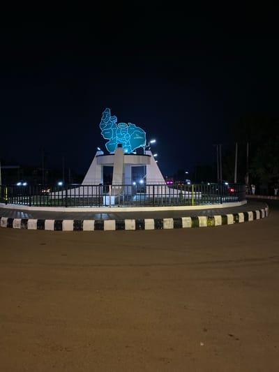 The image shows a night scene at a roundabout in Kuru Dungus, Nigeria. A prominent monument in the center of the roundabout features a large, illuminated map of a region, with several place names visible. The roundabout itself is surrounded by a black metal fence and has a curb painted with alternating black and white stripes. The road surface appears to be asphalt. Several streetlights illuminate the scene, casting a warm glow. In the background, there are trees, utility poles, and some distant buildings. A few figures, possibly pedestrians, can be seen on the right side of the image. The overall atmosphere is that of a well-lit urban space at night.