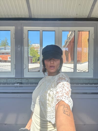 A woman with dark hair, wearing a black cap and a white lace dress, is positioned in front of a window with multiple panes. Reflected in the window are views of buildings, including a brick structure and trees. The background suggests an urban setting. The woman has a tattoo on her arm. The scene appears to be indoors. Given the location hint (Tunbridge Wells, United Kingdom), the architecture and overall environment are consistent with that area.