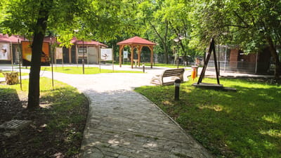 The image captures a tranquil scene in what appears to be a park in the city of Karnobat, Bulgaria. A stone-paved path winds into the frame, flanked by green grass. In the background, there are structures with red-roofed canopies, possibly information kiosks, a gazebo, and caged enclosures. A bench and various posts line the pathway. Trees provide shade, creating a play of light and shadow on the path and grass.