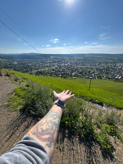 In the foreground of the image, a person's arm with a tattoo of two koi fish is extended, palm open, towards a vast cityscape. A smartwatch is visible on their wrist. The arm is clad in a grey long-sleeved shirt. The scene is set on a grassy hillside overlooking the city of Sim, Russia. The city is densely populated with numerous houses, visible roads, and some industrial structures, including a tall, thin smokestack in the distance. The landscape is characterized by rolling green hills and a clear blue sky with scattered white clouds. Power lines stretch across the frame, originating from the upper left corner and disappearing towards the horizon. The sunlight is bright, casting shadows on the hillside, suggesting a sunny day. The overall impression is one of a scenic overlook, offering a panoramic view of the urban area nestled within a natural landscape.