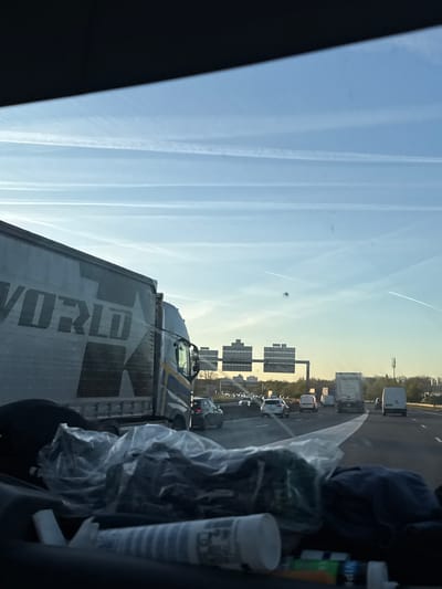 The image shows a view from inside a vehicle traveling on a highway. A large truck with "WORLD" printed on its side is visible in the foreground. In the background, traffic moves on the highway, passing under overhead signs. The sky is light blue with contrails. Other vehicles, including trucks and cars, are visible in the traffic. The exact location cannot be determined from the image, nor can it be conclusively placed in Émerainville, France.