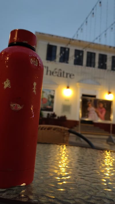 A red water bottle stands in the foreground, partially obscuring a glass table on which reflections of yellow lights are visible. In the background, a building with the word "Théâtre" written across it is illuminated by two warm lights. Several dark windows are visible on the facade of the building. To the right, a framed image displays two figures, one in a pink dress and another in a pink shirt. The scene appears to be outdoors, possibly in the evening, given the ambient light and reflections. The location is identified as Miraflores, Peru, suggesting a public or commercial space like a cafe or a plaza associated with a theater.