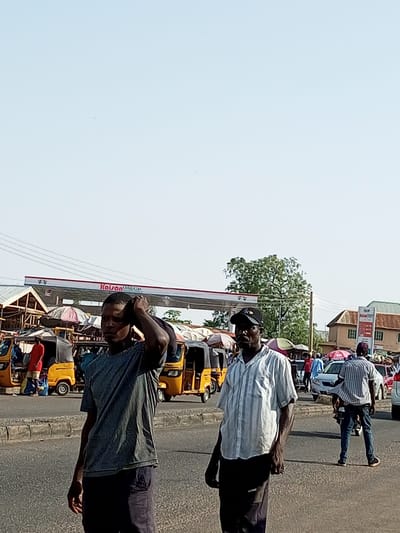 The image shows a street scene in an urban area of Nigeria, likely a market or transportation hub. Two men are walking in the foreground. The man on the left is wearing a dark grey t-shirt and dark pants, with his right hand touching the back of his head. The man on the right is wearing a light-colored, vertically striped short-sleeved shirt and dark pants, with a black baseball cap. Behind them, several yellow auto-rickshaws, commonly known as "keke napep" or "Marwa", are parked or moving. A gas station with a prominent "Kaison Oil & Gas" sign is visible in the mid-ground, along with market stalls and umbrellas. Further in the background, buildings and more vehicles, including a white car and a red car, are present. The sky is clear and pale blue. The scene appears to be during daylight hours.
