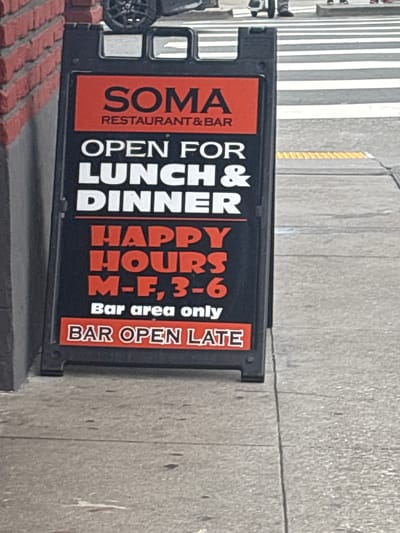 An A-frame sign advertising "SOMA RESTAURANT & BAR" is positioned on a sidewalk in front of a brick building. The sign is primarily black with red and white text. It states "OPEN FOR LUNCH & DINNER" in large white lettering. Below this, in large red lettering, it reads "HAPPY HOURS M-F, 3-6". Further down, in smaller white text, it says "Bar area only". The bottom of the sign has a red section with white text that reads "BAR OPEN LATE". The sidewalk is made of concrete with visible seams. In the background, a crosswalk with white stripes and a yellow tactile paving strip is visible, along with blurred figures of pedestrians and a portion of a vehicle. The overall scene suggests an urban commercial environment.