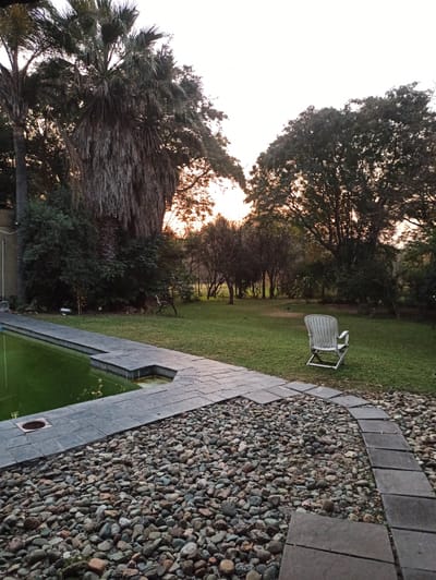 The image depicts an outdoor swimming pool area at dusk, with the sun setting behind a grove of trees. The pool has a shallow end and is filled with green water. A stone pathway and a bed of river rocks surround the pool. A white plastic lawn chair sits on the grassy area next to the pool. The scene is set in a residential garden, likely in Midrand, South Africa, given the palm trees visible in the foreground and the style of landscaping. The lighting suggests it is late afternoon or early evening. There is no visible text in the image.