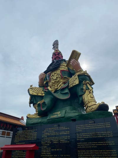 The image shows a large, green and gold statue of Guan Yu, a historical Chinese figure who is revered as a deity. The statue is seated and holds a book in its right hand. It is positioned in front of a dark monument with Chinese and English inscriptions. The monument is located in Baan Hua Thanon, Thailand, as indicated by the text on the monument and the temple architecture visible in the background. The sky is overcast, with some sunlight filtering through the clouds. The statue is the primary subject, depicted in a static, imposing pose. The surrounding environment includes traditional Thai temple structures with red roofs and ornate decorations. The overall scene conveys a sense of reverence and cultural significance.