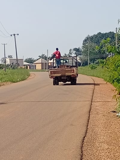 The image shows a man wearing a red shirt and blue jeans standing on the back of a flatbed truck as it drives down a paved road. He is holding a long, dark object, possibly a piece of wood or pipe, over his shoulder. The truck is carrying several bricks. In the background, there are buildings, power lines, and lush green vegetation, consistent with the environment of Du, Nigeria. The sky is clear and bright, indicating a sunny day.