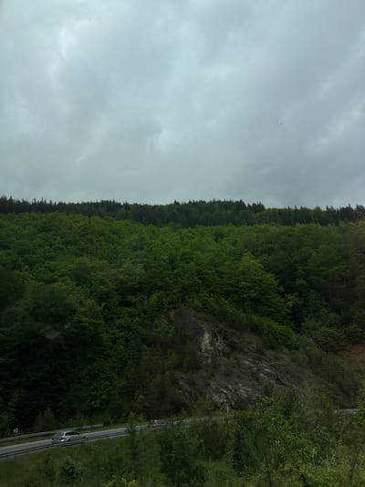 The image depicts a wooded hillside adjacent to a road. The hillside is densely covered in trees, with exposed rock faces visible in some areas. A two-lane road runs along the base of the hillside, with at least two cars visible traveling on it. The sky is overcast with a uniformly gray cloud cover. The image is vertically oriented. There are no people visible in the image. The location is purported to be near Liaskovo, Bulgaria, but this cannot be verified from the image alone.