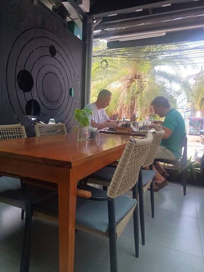 Two men are seated at a wooden table in an outdoor dining area in Ban Ao Nang, Thailand. The man on the left wears a white t-shirt, while the man on the right wears a green t-shirt. Both are focused on eating their meals. The table is set with plates, utensils, and a small plant in a clear vase. Behind the men is a black decorative panel with circular cutouts. Wicker-backed chairs with light blue cushions surround the table. In the background, palm trees are visible along with a street scene that includes buildings and a food cart. The floor is light grey tile. The scene is well-lit, suggesting it is daytime.