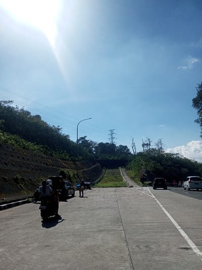 The scene depicts an outdoor roadway on a bright, sunny day in Ka Thaung (Upper), Myanmar. The sky is predominantly clear blue, with the sun producing a strong glare in the upper left quadrant and a few scattered white clouds visible on the right.
The foreground features a paved, multi-lane concrete road. A solid white line delineates the rightmost lane. To the left of the main road is a paved shoulder or access area.
On the far left, a steep, engineered embankment rises, covered in dark soil retention netting or blocks and dense green vegetation. Utility poles with multiple power lines run horizontally across the middle of the frame, parallel to the road and the embankment. A tall, metal lattice power transmission tower is visible on the hillside further back. A street light pole stands near the center-left.
In the mid-ground, on the left shoulder, a dark motorcycle (scooter type) is stopped or parked. Two individuals are on the motorcycle; one wears a light-colored top and a helmet, the other a darker top and a helmet. Behind them, a dark-colored SUV is parked. A person wearing a blue shirt and possibly a hat is standing near the SUV, facing away from the camera, holding what appears to be a bag or container. Another dark vehicle is partially visible further down the shoulder.
To the right of the main road, dense green foliage and trees line the horizon. A gradual grassy incline, with a concrete ramp or steps structure, ascends towards the background. Another power transmission tower is visible in the distance on the right side of the frame, partially obscured by trees.
On the main road's right lanes, a black sedan or SUV is traveling away from the camera. Further behind it, a white minivan or SUV is also in motion, moving away from the camera.