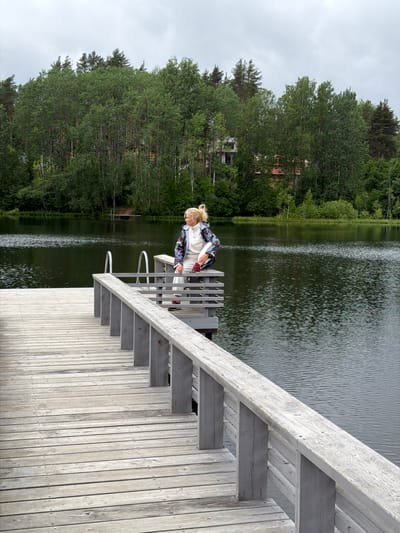 A blonde woman sits on a wooden dock overlooking a lake. She is wearing a white hooded sweatshirt, white pants, and a colorful jacket with a black, white, and blue abstract pattern. Red sneakers are on her feet. The woman is looking to her right, with her legs bent and one arm resting on her knee. The dock is made of weathered grey wood planks and has a wooden railing. In the background, the dark water of the lake is calm, reflecting the overcast sky and the dense green trees that line the opposite shore. The trees are a mix of deciduous and coniferous types, with some houses visible amongst them. The overall scene is serene and natural. The image is not located in Mars, Russia.