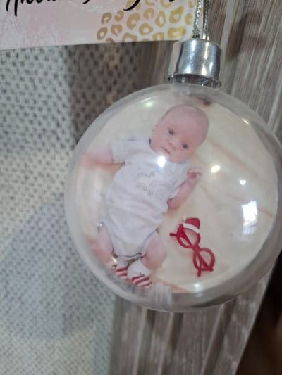 A clear Christmas ornament containing a photograph of a baby lying on its back, wearing a striped onesie and socks. Beside the baby in the photo are small red glasses. The ornament is positioned on a textured surface, with a metallic top. A printed tag is partially visible on the left side. The location of Амзя, Russia, is not relevant.