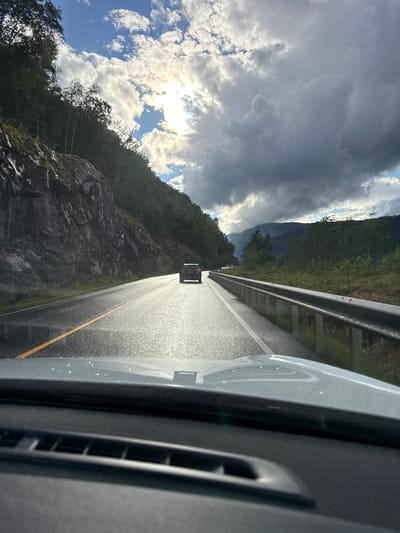 This is a first-person perspective shot from inside a vehicle driving on a wet, asphalt road that curves to the left. The road is marked with a solid yellow line on the left side and a white dashed line on the right. The terrain consists of a steep, rocky, and tree-covered mountainside on the left, and a guardrail bordering a vegetated slope on the right. The sky is partially overcast with dramatic, dark clouds and bright patches of sunlight breaking through. In the distance, more mountains are visible. A black SUV with a bicycle rack is driving in front of the camera vehicle. The scene is set in the natural landscape near Eidfjord, Norway, indicated by the mountainous terrain and road infrastructure characteristic of the region. The wet road surface suggests recent rainfall or mist.