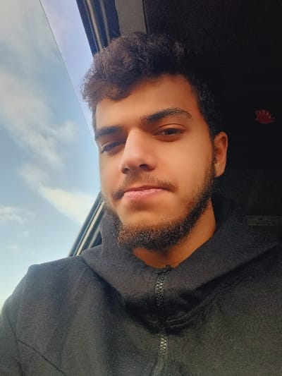 A portrait photograph, taken in Kafr Sousa Municipality, Syria, captures a young male from the chest up, seated inside a vehicle. The subject, likely in his late teens or early twenties, has medium brown skin, dark curly hair, a short beard, and dark eyes. He is wearing a black hooded jacket with a black zipper partially fastened, and looks directly at the camera with a neutral expression. To the subject's left, a window reveals a clear blue sky with sparse white clouds. To his right, the dark interior of the vehicle is visible, with a small, blurry red object positioned in the upper right corner. No other individuals, objects, or actions are discernible within the frame.