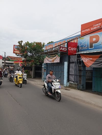 The image shows a street scene in Kecamatan Kutawaringin, Indonesia (latitude: -6.974, longitude: 107.542). Six people are riding motorbikes on a paved road, traveling from left to right.  A white motorbike with a blue license plate is in the foreground, with a man wearing a blue shirt and a black cap riding it.  The man is wearing a blue shirt and a black cap. The road is flanked by sidewalks and buildings. There are several shops with signs in the background.  The shops have blue and white walls and black metal gates.   There is a green tree in the left background.  The sky is overcast and gray.  The scene is in daytime.  The image was likely taken on a street near a commercial district.