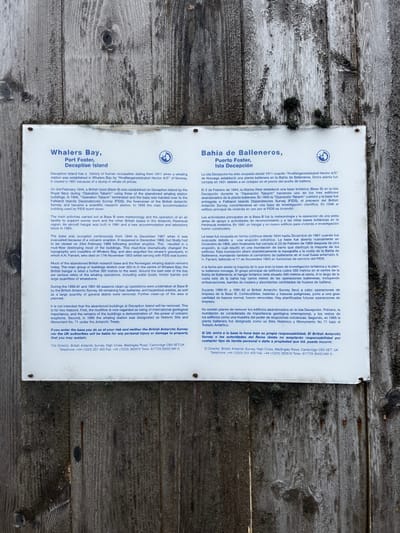 The informational sign details the history of Whalers Bay, Port Foster, and Deception Island. It mentions the Norwegian whaling station established in 1911, its closure in 1931, and the British base (Base B) established during "Operation Tabarin" in 1944. The sign describes the activities, volcanic eruptions that impacted the base, and clean-up operations. It explains why the abandoned buildings remain and warns visitors about entering at their own risk. Contact information for the British Antarctic Survey is provided. The same information is written in Spanish on the other half of the sign.