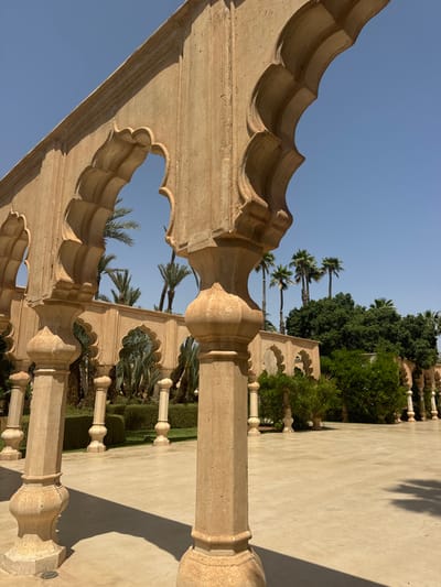 The image shows an arched walkway lined with stone pillars in a courtyard in the city of Ouled Jelal, Morocco. The pillars are all made of a light tan stone and are adorned with intricate carvings. The walkway leads to a paved courtyard. There are palm trees and other greenery lining the courtyard and the walkway. The sky is blue and cloudless. The image was taken from the perspective of someone standing under one of the arches, looking toward the courtyard. The image was taken at a location with coordinates latitude: 31.663, longitude: -7.872.