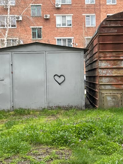 The image shows a gray metal garage door with a spray-painted heart outline, positioned between a weathered train car and a brick building. The location is Dmitrievskaya, Russia, indicated by the presence of Russian architectural elements. The train car's underside overhangs the garage. Grass and weeds grow along the garage's right side. The brick building has multiple windows and air conditioning units. There are bare tree branches visible in front of the brick building. The photograph appears to be taken at ground level.
