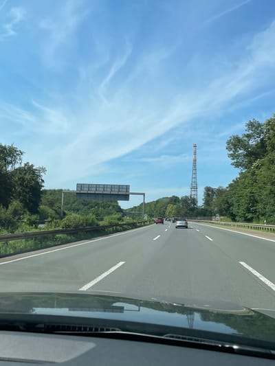 The image captures a view from the perspective of a vehicle traveling on a highway. The highway has multiple lanes marked with white lines, and several cars are visible in the distance. On the right side of the highway, dense green trees line the road, and a tall, metallic tower with red and white accents stands prominently against the blue sky with wispy white clouds. On the left side of the highway, there is a large overhead sign structure, also surrounded by trees and greenery. The foreground of the image shows the dashboard and windshield reflection of the car, including a distorted reflection of the tower and trees. The scene appears to be on a clear, sunny day. Based on the context of Langenfeld (Rheinland), Germany, this highway is likely part of the Autobahn network.