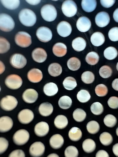 The image shows a close-up of a black perforated surface with round holes. The holes reveal a blurry, out-of-focus background with various colors including white, gray, orange, and hints of blue and green. The specific location in Charentsavan, Armenia, is not discernible from the image. There are no people, actions, or interactions visible.