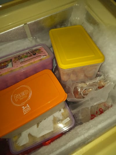 This image depicts the interior of a domestic freezer in Tarqumiyah, Palestinian Territory, characterized by visible frost buildup on the inner walls. Several food items are stored within various containers and plastic bags.
In the upper right, a rectangular clear plastic container with a yellow lid holds a quantity of reddish-brown, possibly frozen, food product. To its left, another clear rectangular container with a pink-purple lid, decorated with cartoon characters, contains packaged items, including an "Indomie" branded product.
In the lower left, an orange-lidded rectangular container, branded "NEO COFFEE 3 IN 1 INSTANT COFFEE," rests on top of a clear plastic container with a light purple base, which holds a light-colored substance partially covered by white paper or tissue. Transparent plastic bags containing various food items, including what appears to be reddish-brown meat, are visible on the right side, nestled among the containers and against the frosted freezer wall. No individuals are present, and no actions are being performed.