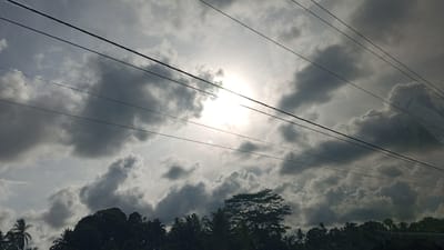The image depicts a cloudy sky with the sun partially obscured by dense clouds. Several power lines traverse the frame horizontally. In the foreground, the silhouettes of lush green trees and palm trees are visible, suggesting a tropical or subtropical environment. The location is identified as Luengbaro, Indonesia. There are no people or discernible actions in the scene. The overall impression is one of overcast weather with the sun attempting to break through.
